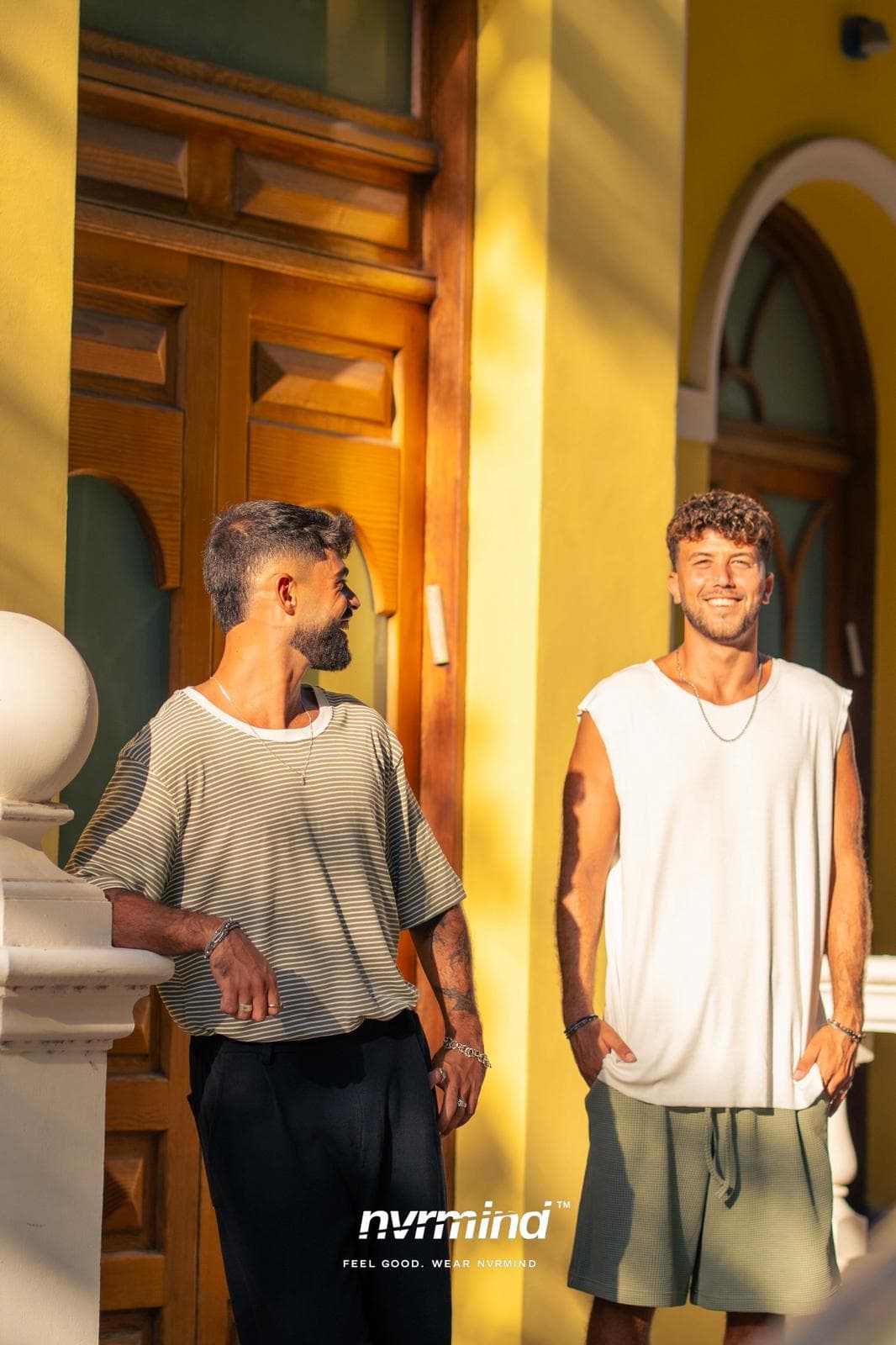 Two men in NVRMIND oversized fits, laughing together at a sunlit doorway.